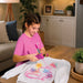 A young girl with braided hair sits at a wooden table in a cozy living room, using a pink spray paint pen to draw colorful butterflies and flowers on a white sweatshirt.