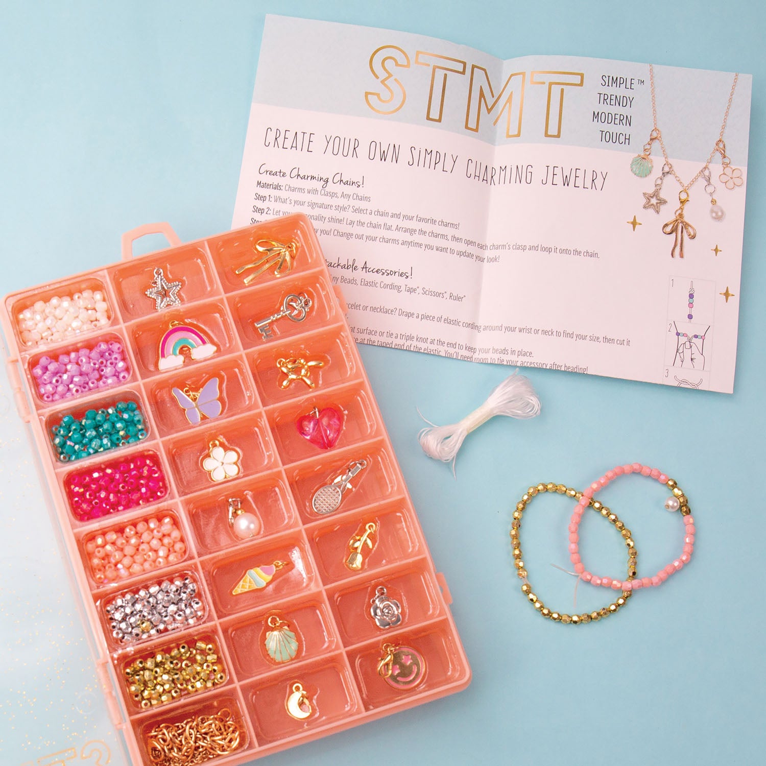 A STMT jewelry-making kit laid out on a light blue background, including a peach organizer filled with beads and charms, an instruction booklet, thread, and two finished beaded bracelets.