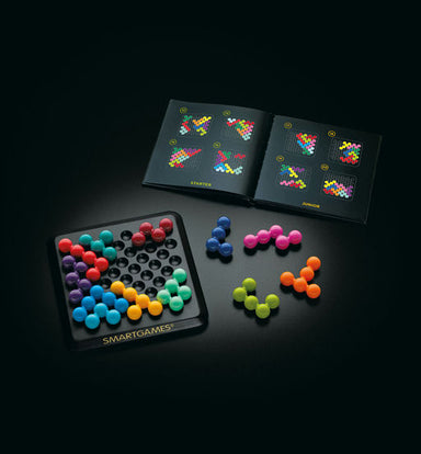 A board game set displayed on a dark surface, featuring a black game board with colorful pegs, several loose colorful pegs, an open instruction booklet, and a branded box. The visible brand on the product is SMARTGAMES.