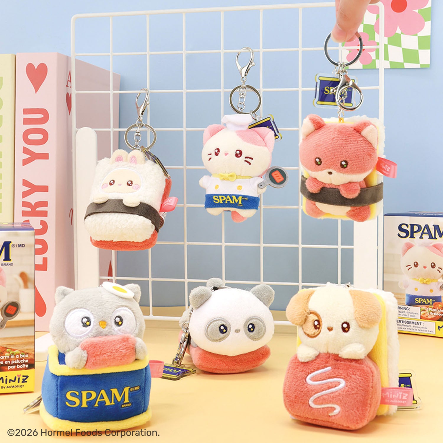 A group of cute plush character keychains and small pouches displayed on a tabletop with a white grid rack behind them. The characters include a white cat in a sushi roll, a pink fox in a sushi roll, a gray owl in a sandwich, a panda, and a dog in a sandwich.