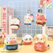 A group of cute plush character keychains and small pouches displayed on a tabletop with a white grid rack behind them. The characters include a white cat in a sushi roll, a pink fox in a sushi roll, a gray owl in a sandwich, a panda, and a dog in a sandwich.