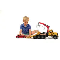 A young boy in a blue shirt sits on a yellow toy car carrier truck with a crane, playing on a white studio background.