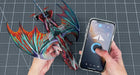 A 3D-printed articulated dragon model with vibrant multicolor wings (red, orange, teal, purple) is held by two hands over a gray gridded cutting mat, next to a smartphone displaying a control app.
