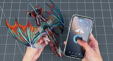 A 3D-printed articulated dragon model with vibrant multicolor wings (red, orange, teal, purple) is held by two hands over a gray gridded cutting mat, next to a smartphone displaying a control app.
