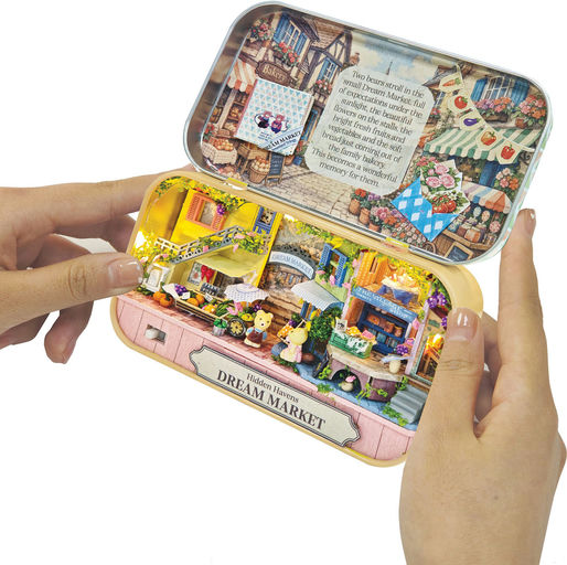 A person's hands holding open a small metal tin-shaped craft box labeled 'Dream Market' and 'Halloween Fair.' The inside features colorful miniature market stalls, tiny figures, a printed story panel on the lid, and warm LED lighting.