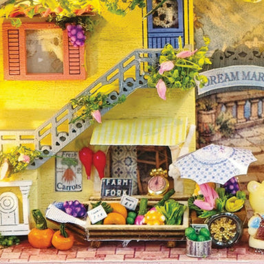 A miniature dollhouse-style village scene with a yellow house, staircase, small market stall, produce cart, flowers, and tiny decorative figures. The scene appears to be the product itself rather than a real environment, so this fits Scenery rather than Lifestyle or Product/Packaging.