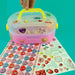 A pink and clear plastic sticker craft kit with a yellow handle, shown with a hand lifting the lid and a sheet of colorful heart-shaped stickers spread out below on a bright teal background.