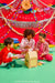 Three children sitting on a green floor in front of a decorated red backdrop, playing with a small wooden toy money box labeled 'MALL'. The scene is colorful and festive, with paper fans, tinsel, and pillows in the background.