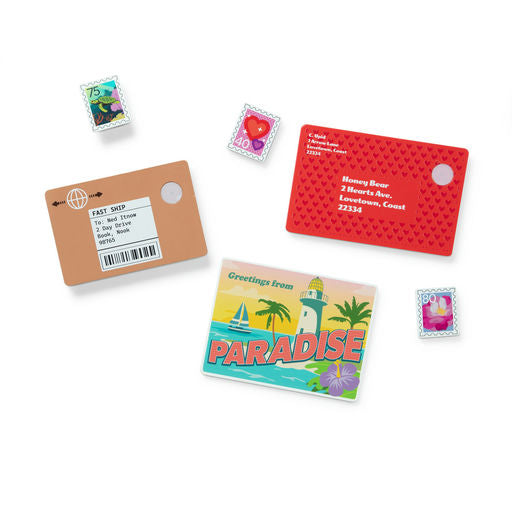 A flat-lay arrangement of postage stamp-themed stationery items on a white background, including a brown shipping card, a red address card with a heart pattern, a colorful 'Greetings from Paradise' postcard, and several small decorative stamps.