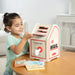 A young girl with dark hair in a mint green shirt is sitting at a wooden table, inserting a card into a Melissa & Doug children's toy mailbox. The toy mailbox is cream-colored with red and black details, and several toy letters and envelopes are placed in front of it on the table.