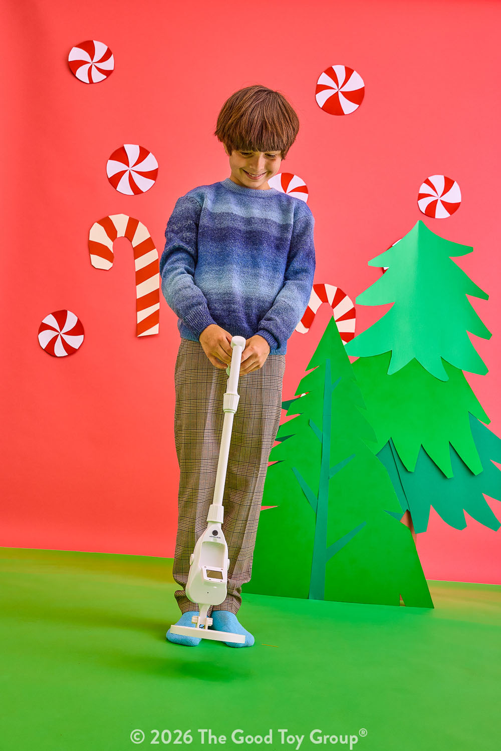 A child stands in a colorful studio setting holding a metal detector. The background is bright red with paper candy canes, peppermint candies, and a green paper Christmas tree.