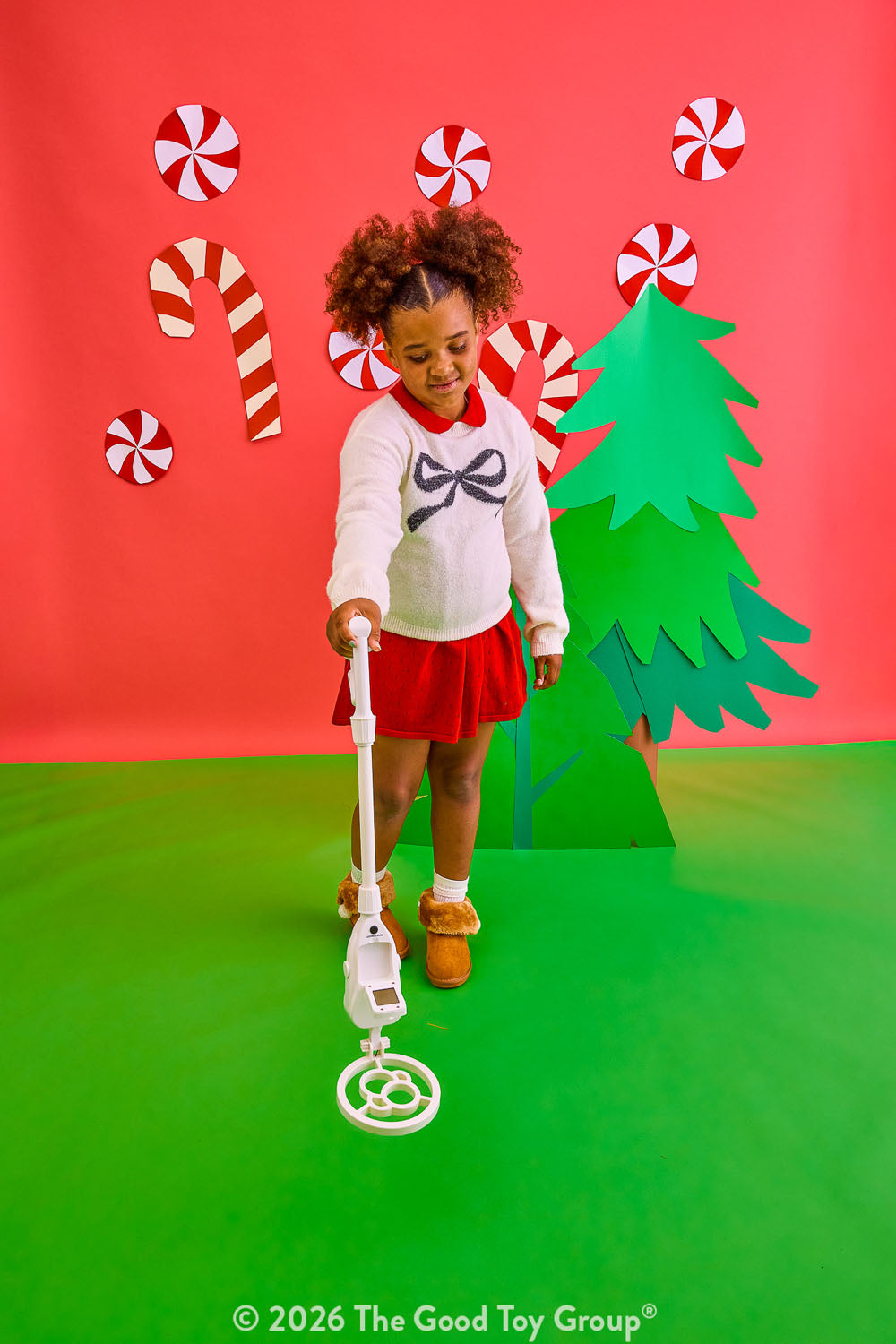 A young child stands on a green floor against a red backdrop, using a metal detector. The scene includes a green paper Christmas tree, red-and-white candy canes, and peppermint candies, with a visible The Good Toy Group watermark at the bottom.