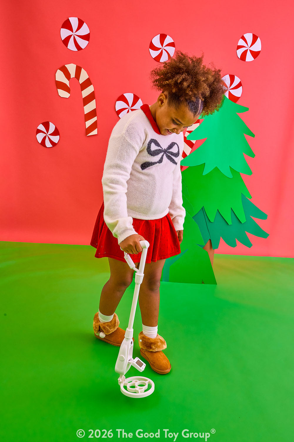 A young child stands on a green floor against a red backdrop, using a metal detector. The child wears a white sweater with a dark bow, a red skirt, and brown shoes.