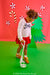 A young child stands on a green floor against a red backdrop, using a metal detector. The child wears a white sweater with a dark bow, a red skirt, and brown shoes.