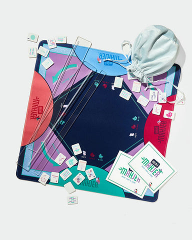A colorful board game set laid out on a light gray background, including a large round board with geometric sections, multiple transparent acrylic game pieces, small white cards, two printed instruction cards, and a light blue drawstring bag.