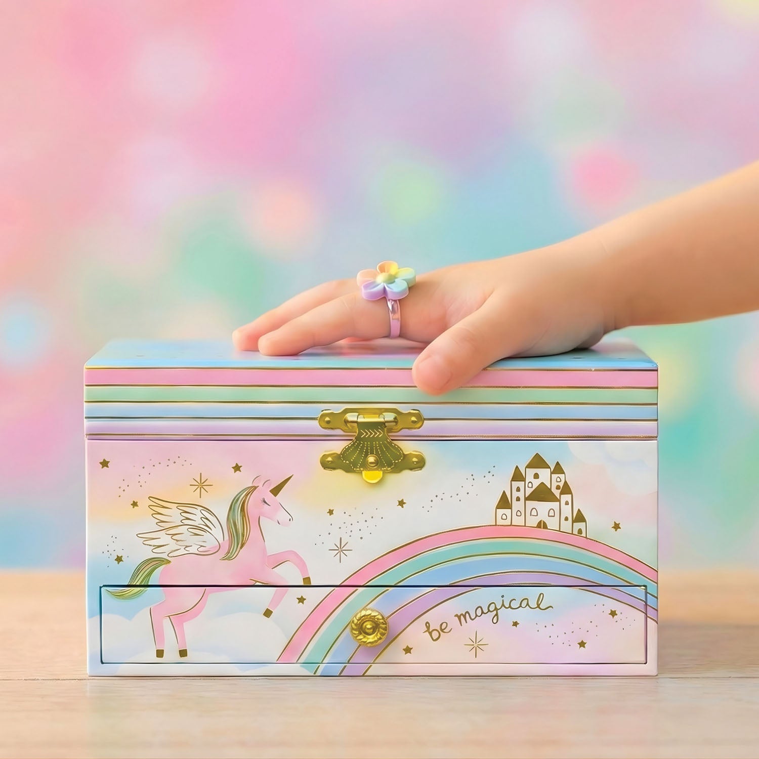 A pastel unicorn-themed jewelry box with a small drawer, decorated with a pink winged unicorn, rainbow, castle, and gold stars. A child's hand wearing a pastel ring rests on top of the box, which is set against a soft, colorful bokeh background.