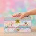 A pastel unicorn-themed jewelry box with a small drawer, decorated with a pink winged unicorn, rainbow, castle, and gold stars. A child's hand wearing a pastel ring rests on top of the box, which is set against a soft, colorful bokeh background.