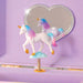 A small white unicorn figurine with a pastel pink, blue, and purple mane and tail, standing on a cloud-shaped base attached to a spring-loaded stand. The unicorn is photographed inside a lavender box or case, with a heart-shaped mirror reflecting the back of the product.
