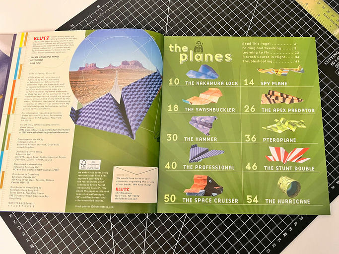 An open Klutz 'the planes' origami booklet spread on a black gridded cutting mat, showing various plane folding projects and instructions.