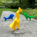 Three colorful origami bird sculptures displayed outdoors on a stone pathway, with green foliage in the background. The birds are in yellow, blue, and green, and the text 'PICKAGRAM - THREE AMIGOS' and 'Pickagram' are visible.