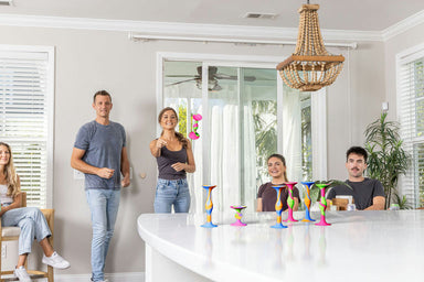 A group of five people in casual home attire gathered around a white kitchen island in a bright, modern room. Several colorful, hourglass-shaped decorative candle holders are arranged on the countertop.