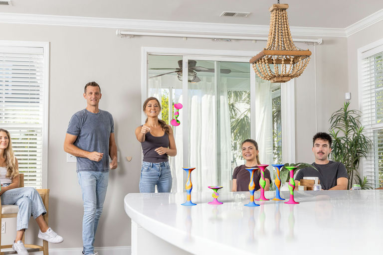 A group of five people in casual home attire gathered around a white kitchen island in a bright, modern room. Several colorful, hourglass-shaped decorative candle holders are arranged on the countertop.