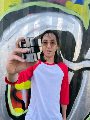 A man with long dark hair and glasses holds up a black and white 3D puzzle cube in front of a colorful graffiti wall. He is wearing a white and red raglan baseball t-shirt.