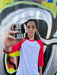 A man with long dark hair and glasses holds up a black and white 3D puzzle cube in front of a colorful graffiti wall. He is wearing a white and red raglan baseball t-shirt.