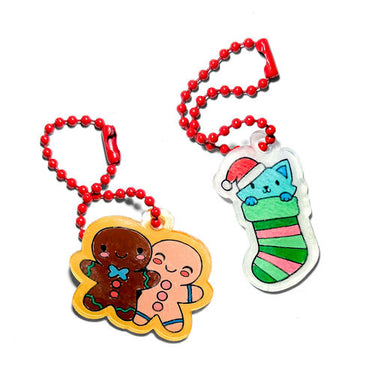 Two Christmas-themed acrylic keychains on red ball chains. One keychain features a cute gingerbread man and gingerbread woman figure with a yellow border.