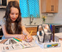 A young girl with long brown hair sits at a kitchen table, writing on a worksheet while using a Spin Animals educational toy. The toy features a clear acrylic cage holding colorful animal figures on a white base with a blue and yellow logo.