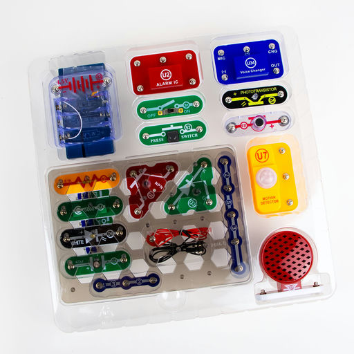 A children's robotics and electronics kit in a clear plastic clamshell package, featuring colorful electronic components such as a blue speaker, red and green buttons, a yellow motor driver, and various connectors on a white background.