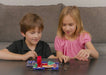 Two children, a boy in a black shirt and a girl in a pink top, sitting on a couch and playing with a small electronic circuit board on a table.
