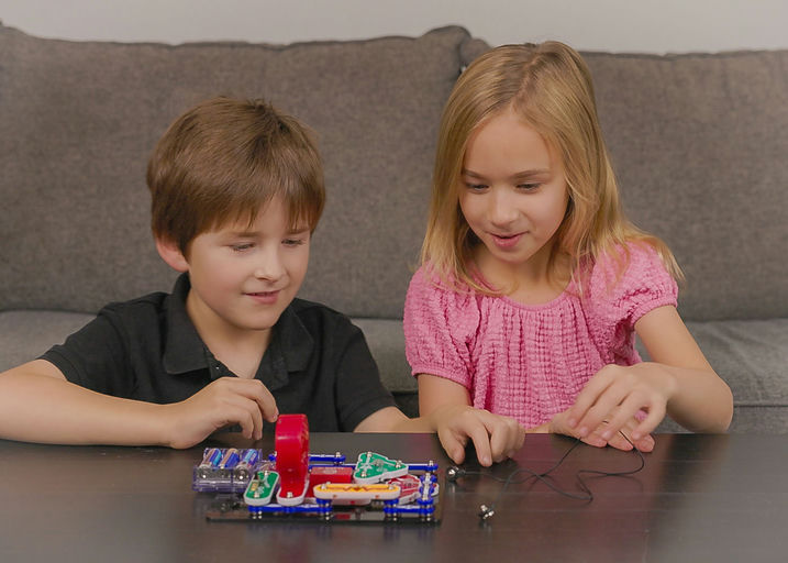 Two children, a boy in a black shirt and a girl in a pink top, sitting on a couch and playing with a small electronic circuit board on a table.