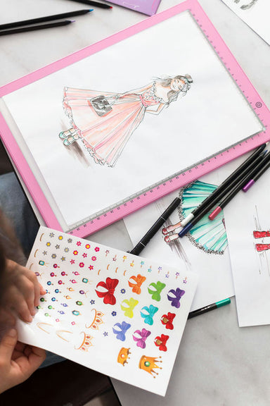 Colorful hand-drawn designs on a white sheet with a pink border, surrounded by art supplies.
