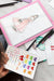Colorful hand-drawn designs on a white sheet with a pink border, surrounded by art supplies.