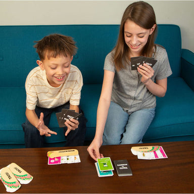 Two children sitting on a teal couch playing a card game at a wooden table, holding cards from the game NOPEK.