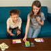 Two children sitting on a teal couch playing a card game at a wooden table, holding cards from the game NOPEK.