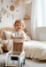A toddler with blonde curly hair stands in a softly lit nursery, smiling while pushing a wooden toy bus with a blue handle. The room has cream bedding, a light wood headboard, and soft neutral decor.