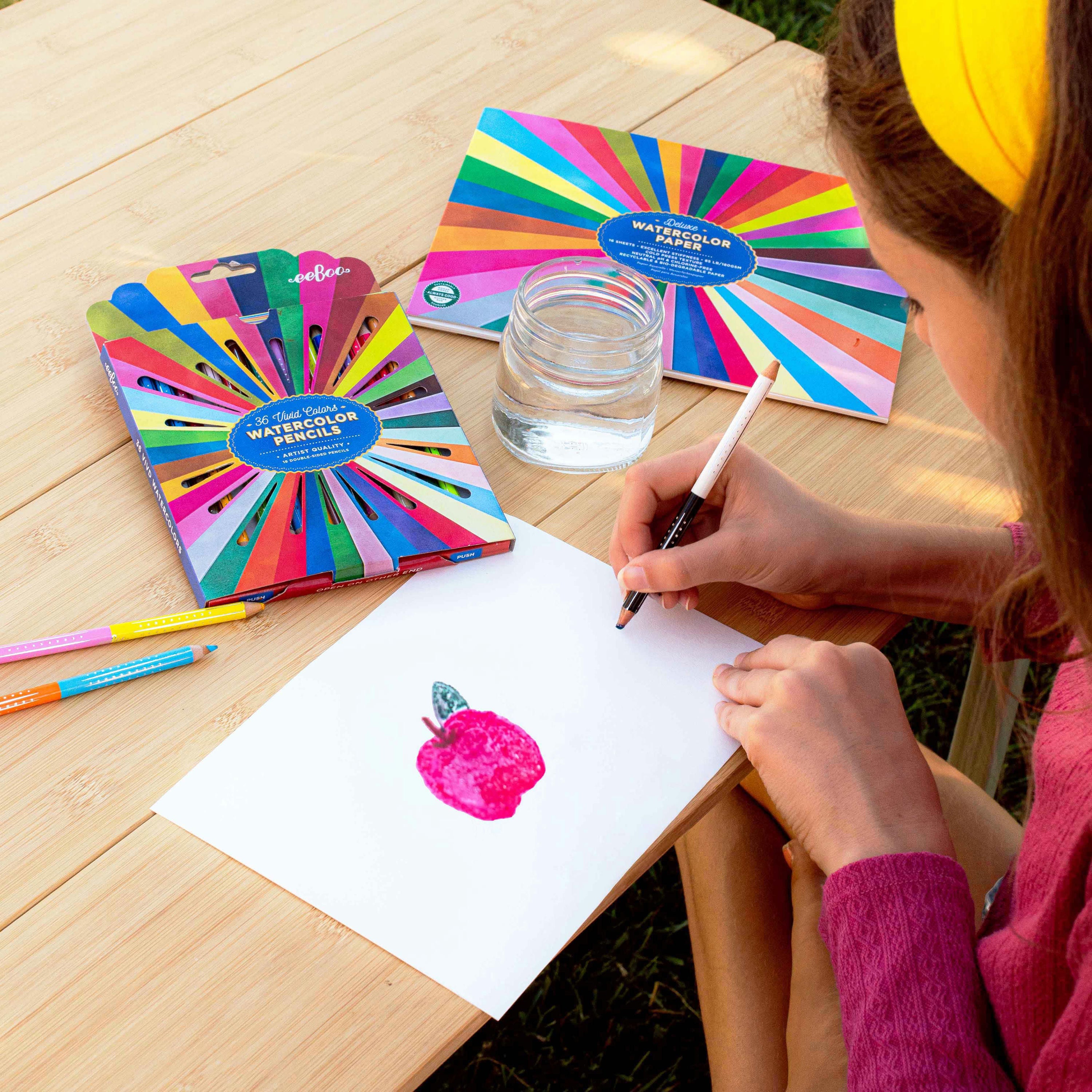 Person drawing with watercolor pencils on a wooden table with a colorful pencil set and water glass.
